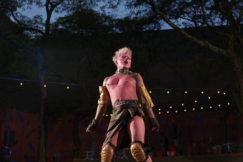 Performance art photograph of a light pink skinned person with spiky blonde hair and dark eye makeup, wearing a studded collar and brown kilt, standing outdoors under string lights.