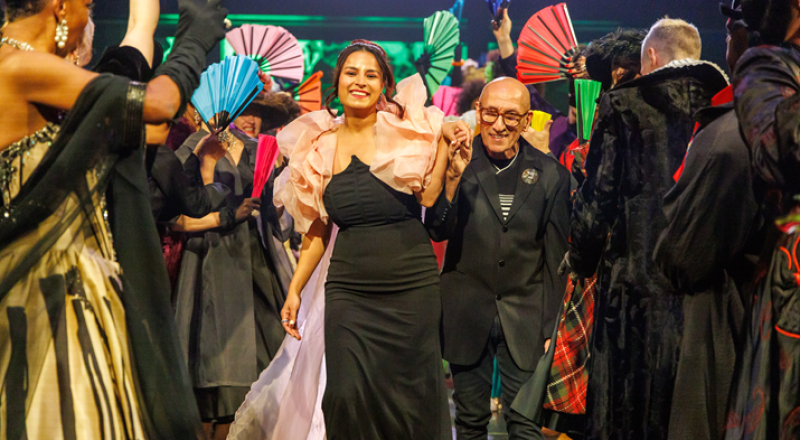 Smiling woman in a black dress with a voluminous pink top, holding hands with a bald man in a black suit, surrounded by people with colorful fans.