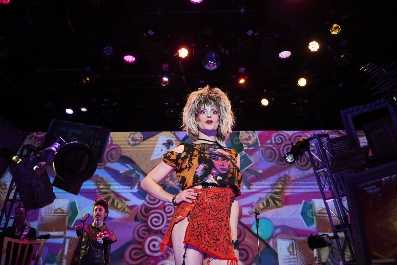 A drag queen with a blonde wig, bold makeup, and a ripped orange and black outfit stands on a stage with colorful lights.