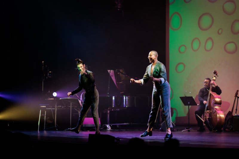 Two women in jumpsuits dance on a dark stage with a green screen, a pianist, and a bassist.