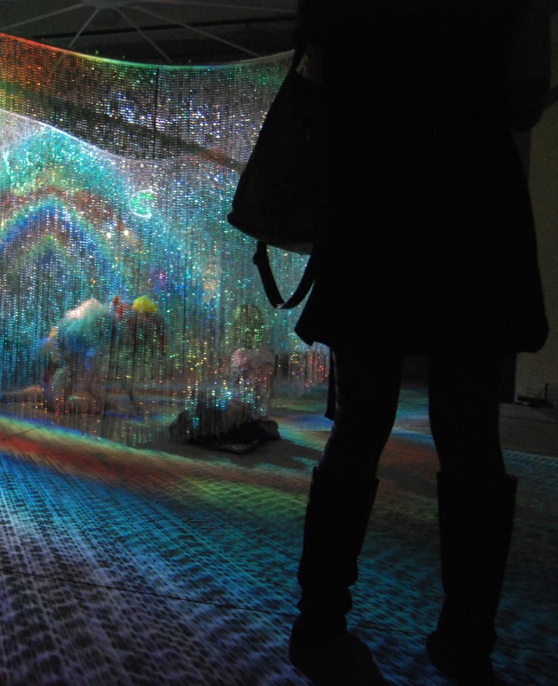 Silhouette of a person standing before a curved, shimmering wall of lights, casting colorful reflections on the floor.