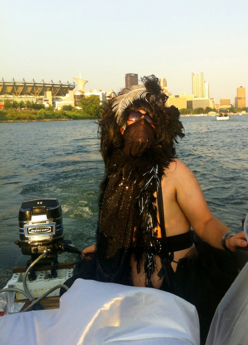 Person in a feathered costume with a mesh mask and sunglasses on a boat, facing away from a city skyline.