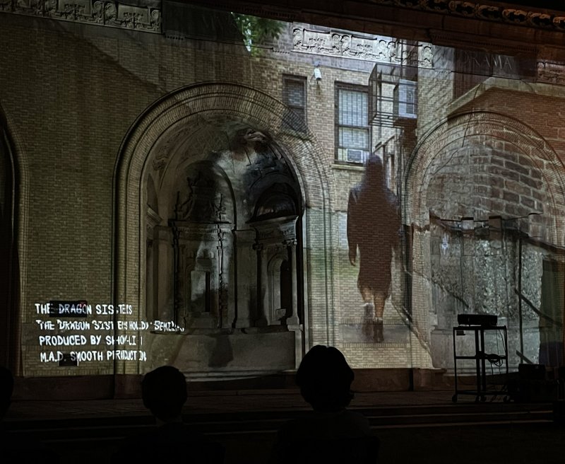 Video projection of a dark figure walking on a brick building with arched windows, text overlay, and two silhouetted viewers.