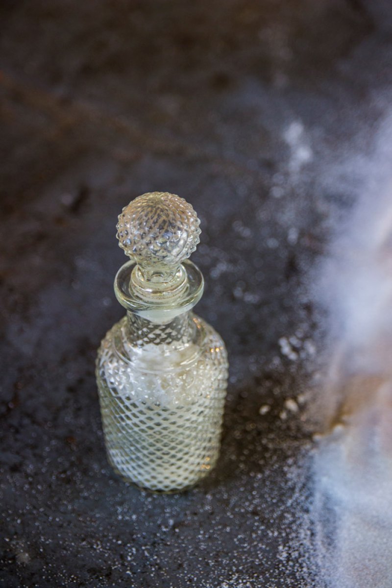 Clear glass decanter with a diamond pattern and spherical stopper, filled with white powder, on a dark, textured surface.