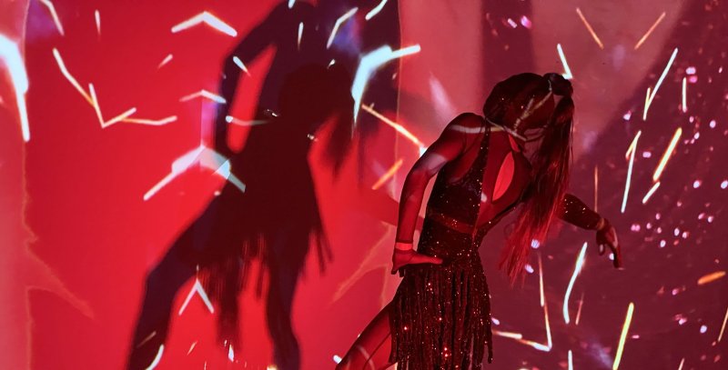 Performer in a sparkly dress with long hair, bending backward, illuminated by red light and white streaks, casting a large shadow.