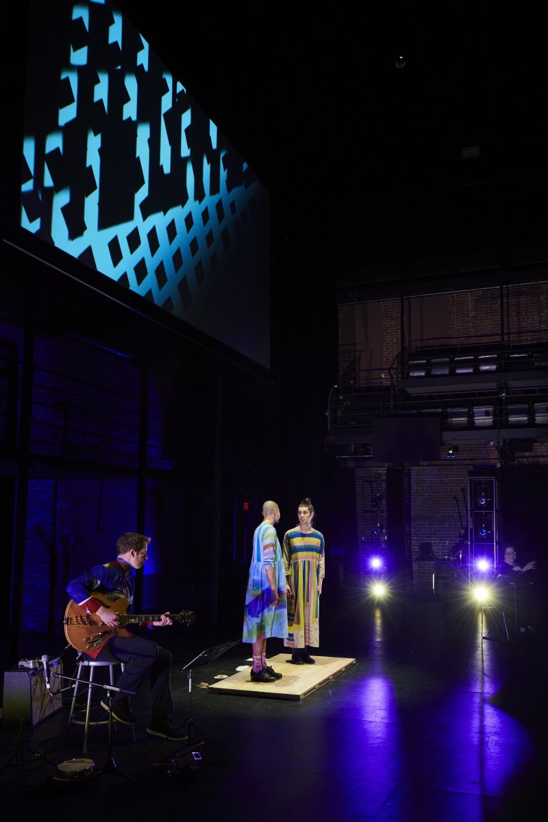 Theater stage view with two figures in colorful dresses on a platform, a guitarist to the left, and a screen projecting blue geometric shapes in the background. Stage lights illuminate the scene.