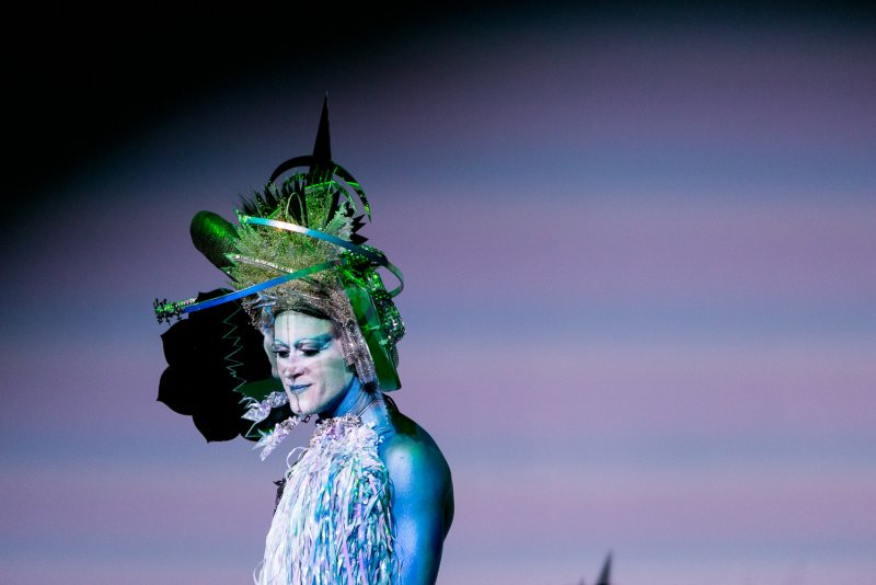Drag performer Darrell Thorne in blue body paint, elaborate green headdress, and white feathered costume against a purple background.