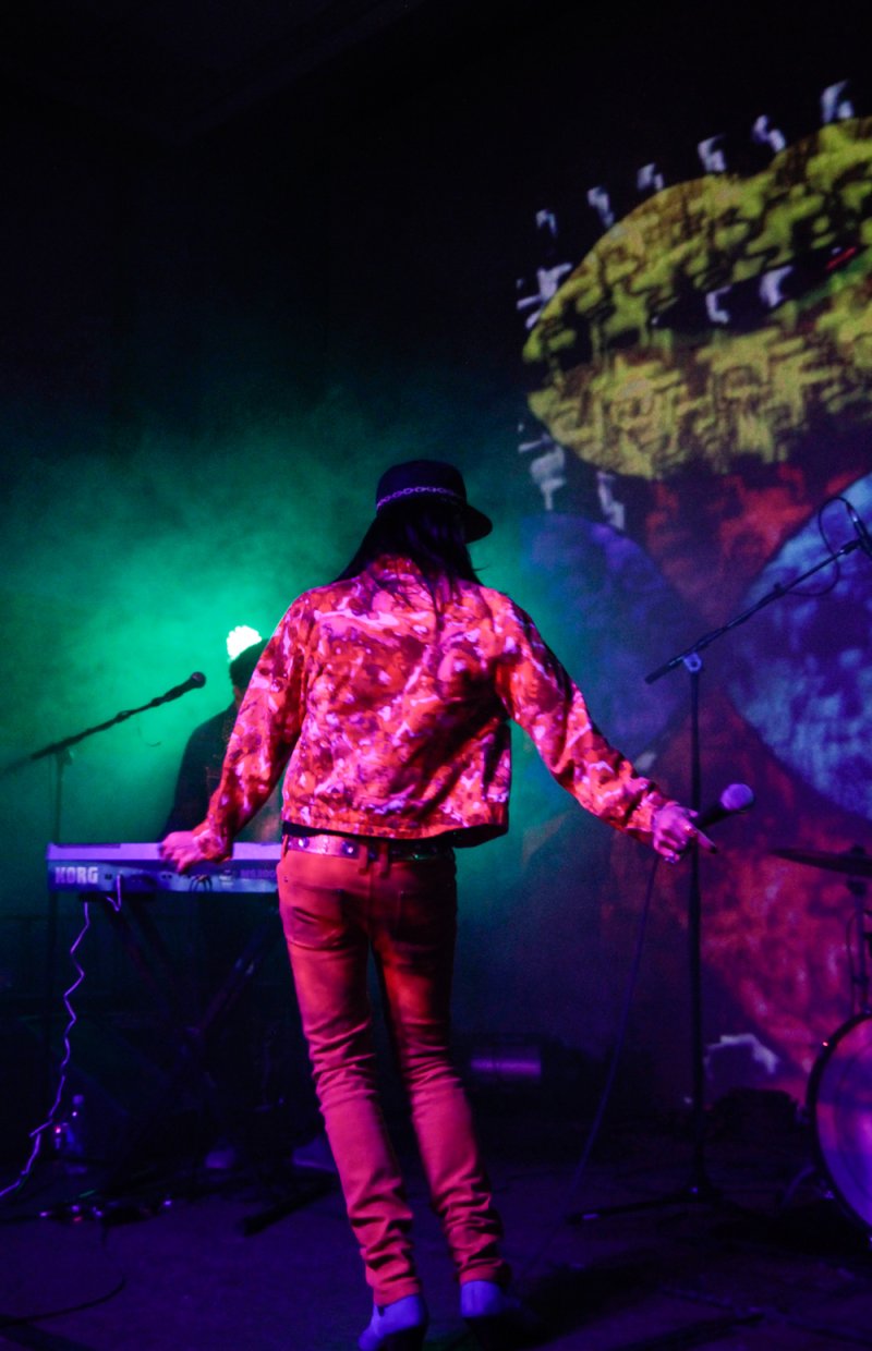 Performer in a vibrant orange jacket and pants, facing away from the viewer, on a stage bathed in green and purple light.