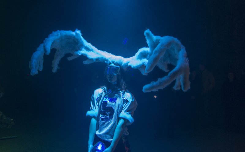 Performer in goggles and white jersey, with large, textured white antlers, illuminated by blue light in a dark space.