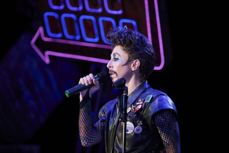 Scott N Andrew as Hedwig, with dramatic makeup, fishnet sleeves, and a leather vest, sings into a microphone on a dark stage with neon lights.