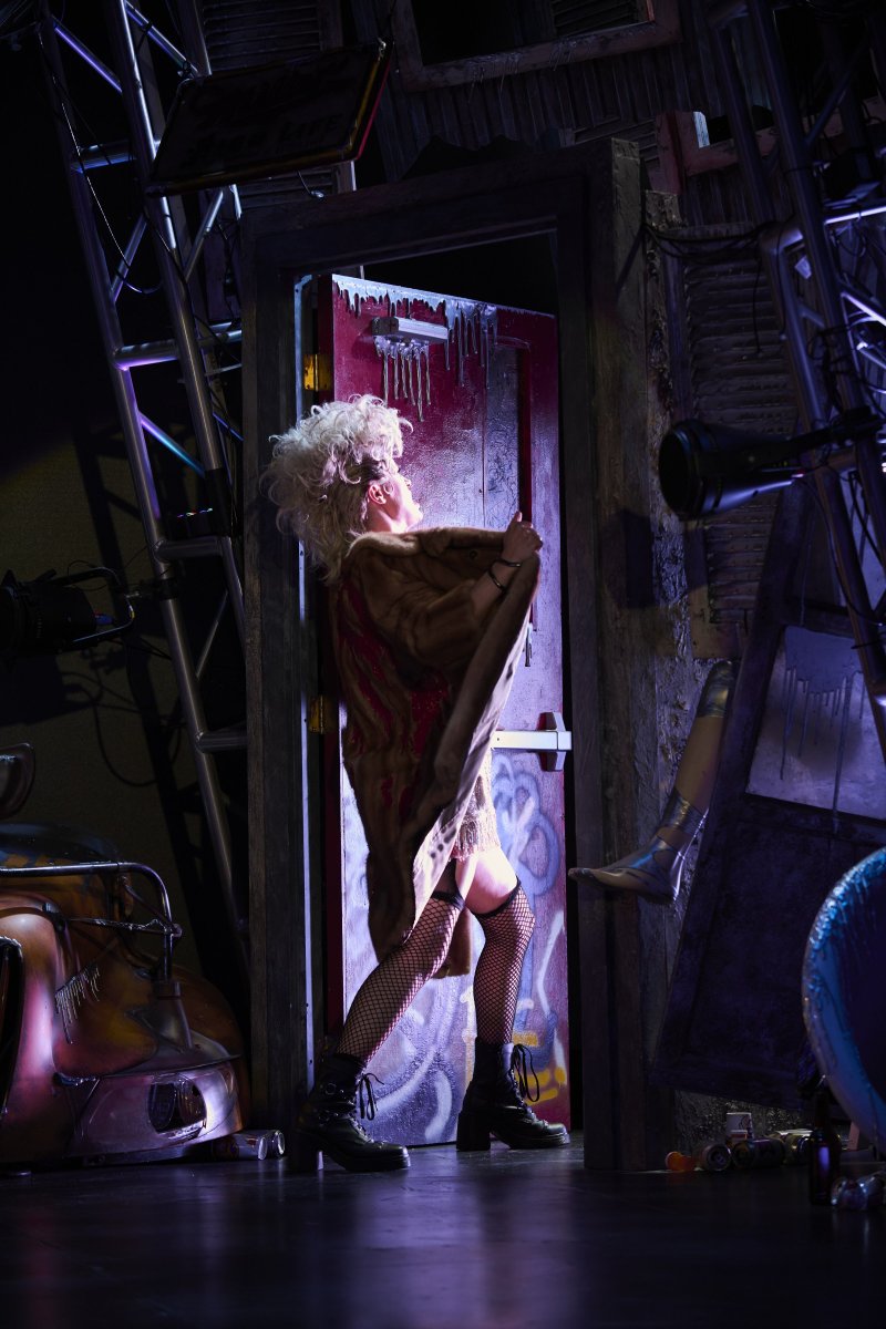 Drag queen in blonde wig, fur coat, fishnets, and boots stands in a doorway with graffiti, surrounded by dark, industrial clutter.
