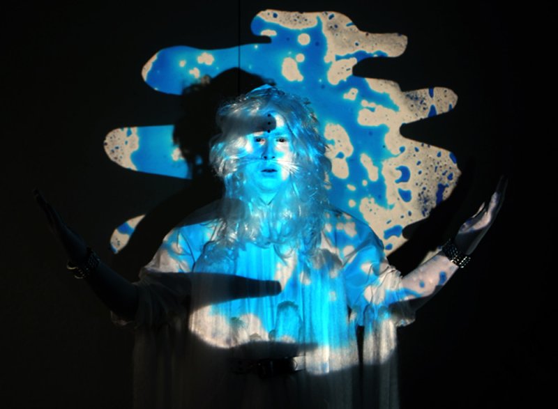 Performance art piece with a person in white wig and robe, illuminated by blue light and a projected blue and white abstract shape on a dark background.