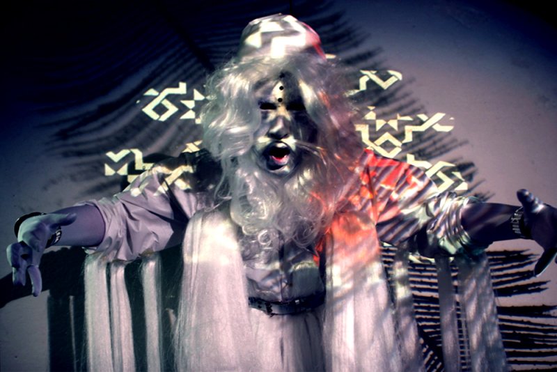 Figure with white wig, silver face paint, and open mouth, arms outstretched. Geometric and palm frond patterns are projected onto the figure and background.