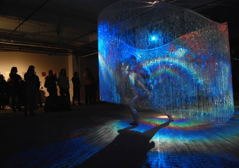 A person stands inside a large, curved curtain of shimmering beads, illuminated by vibrant blue and rainbow lights.