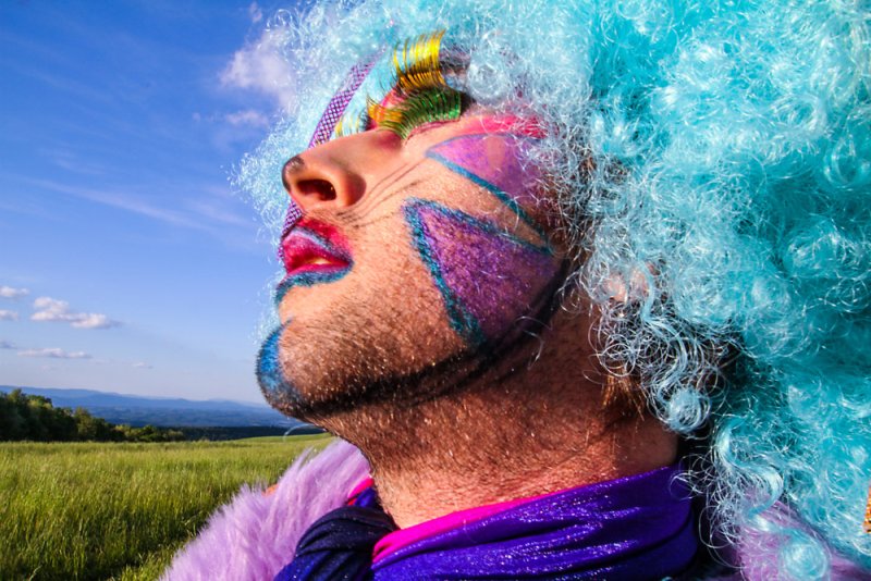 Close-up of a person in drag with bright blue curly hair, colorful face paint, and glittery makeup, looking up at a blue sky.