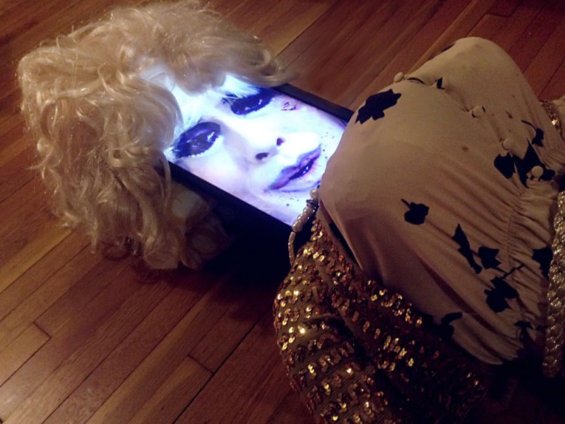 Blonde wig on a screen displaying a woman's face with heavy makeup, attached to a sequined garment on a wood floor.