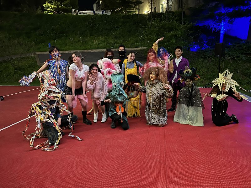 Group of people in elaborate, colorful costumes on a red court at night, with trees and buildings in the background.