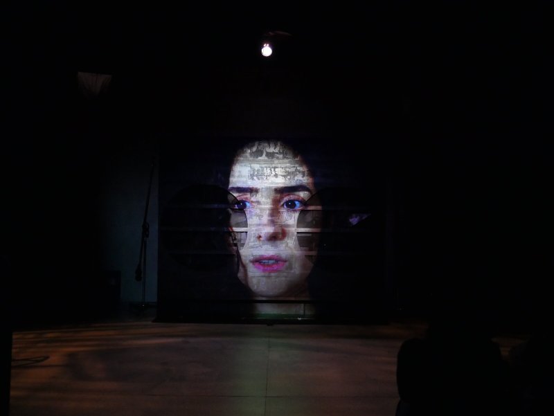 Projected image of a woman's face with dark hair, looking forward, on a screen in a dark room.