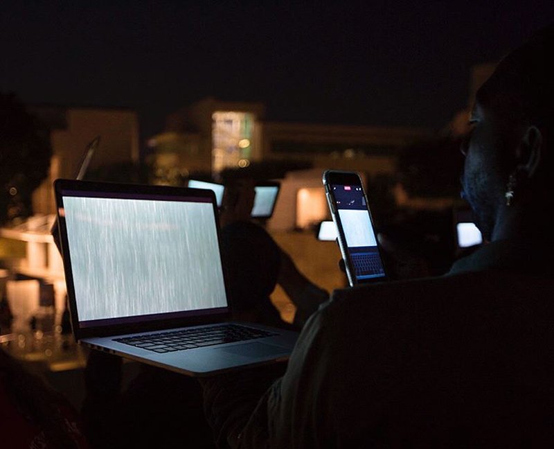 Person in dark watching laptop with static screen and phone displaying a white rectangle, other screens glow in background at night.