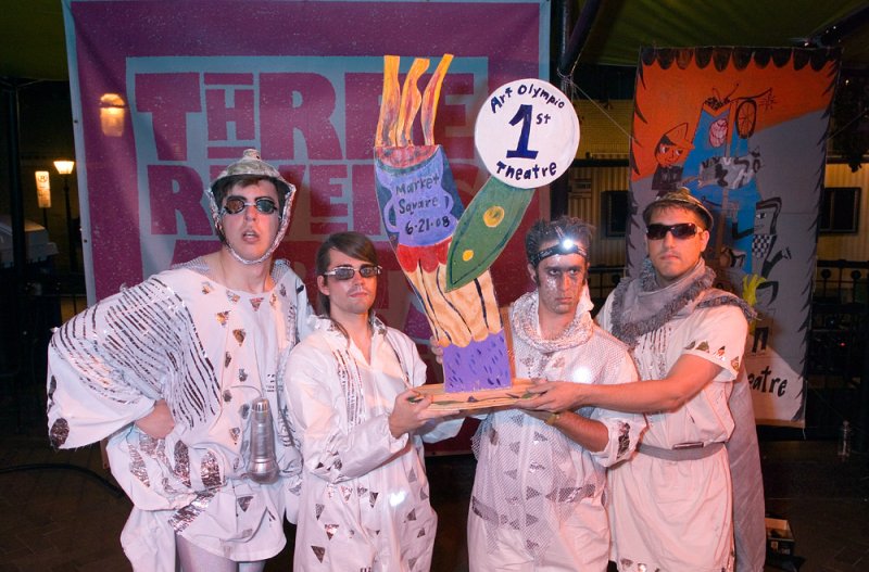 Four performers in white costumes hold a painted trophy in front of colorful backdrops. Costumes are adorned with silver accents. One wears a tin foil helmet and goggles.
