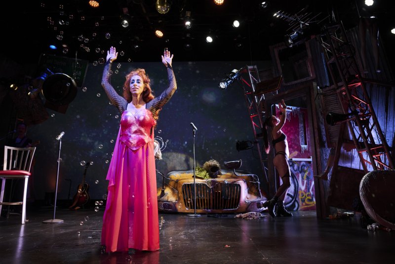 Drag queen in a pink dress and fishnet sleeves on stage, arms raised, with a second performer and a car prop.