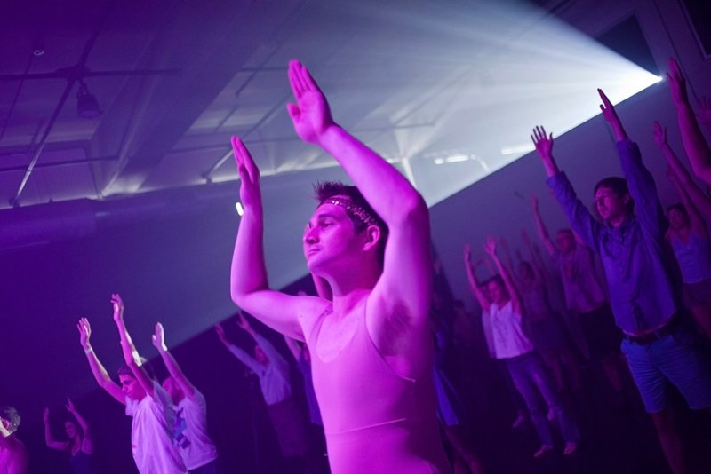 A group of people with their arms raised, illuminated by purple and white stage lights.