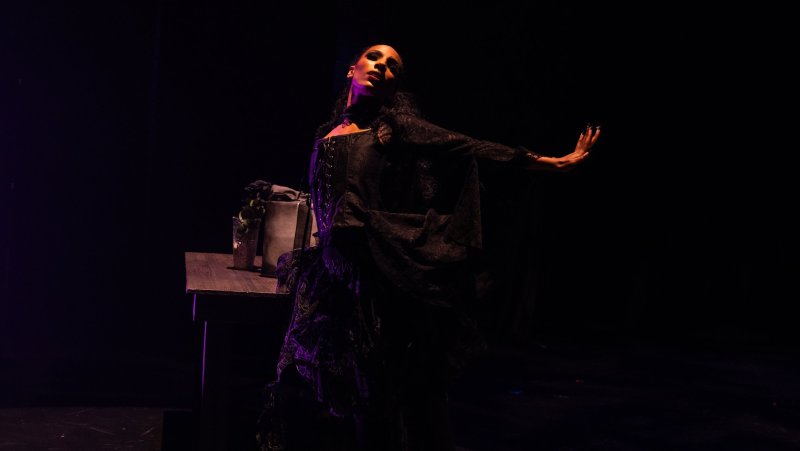 Dark stage scene with a figure in a lace dress and corset, arm outstretched, next to a table with a vase. Purple lighting.