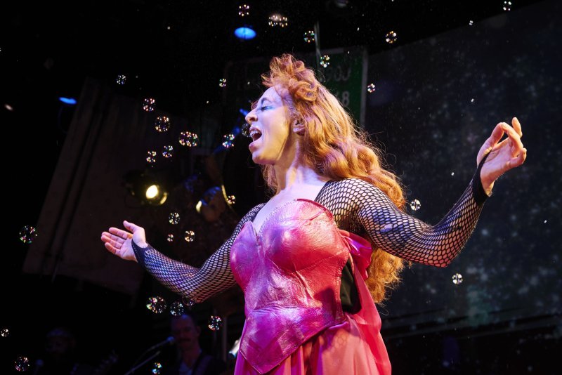 A person with red hair in a pink outfit and fishnet sleeves sings on stage, surrounded by bubbles and stage lights.
