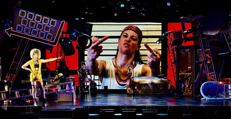 Stage set with a blonde performer in a yellow dress on the left, and a large screen behind showing a person flipping two middle fingers.