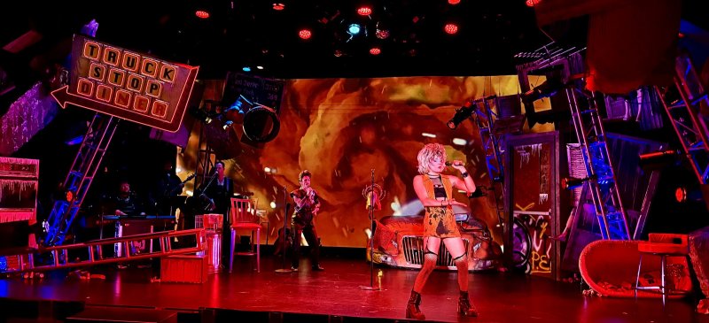 A blonde punk singer in a short dress and fishnets performs on a stage with a "Truck Stop Diner" sign and fiery backdrop.
