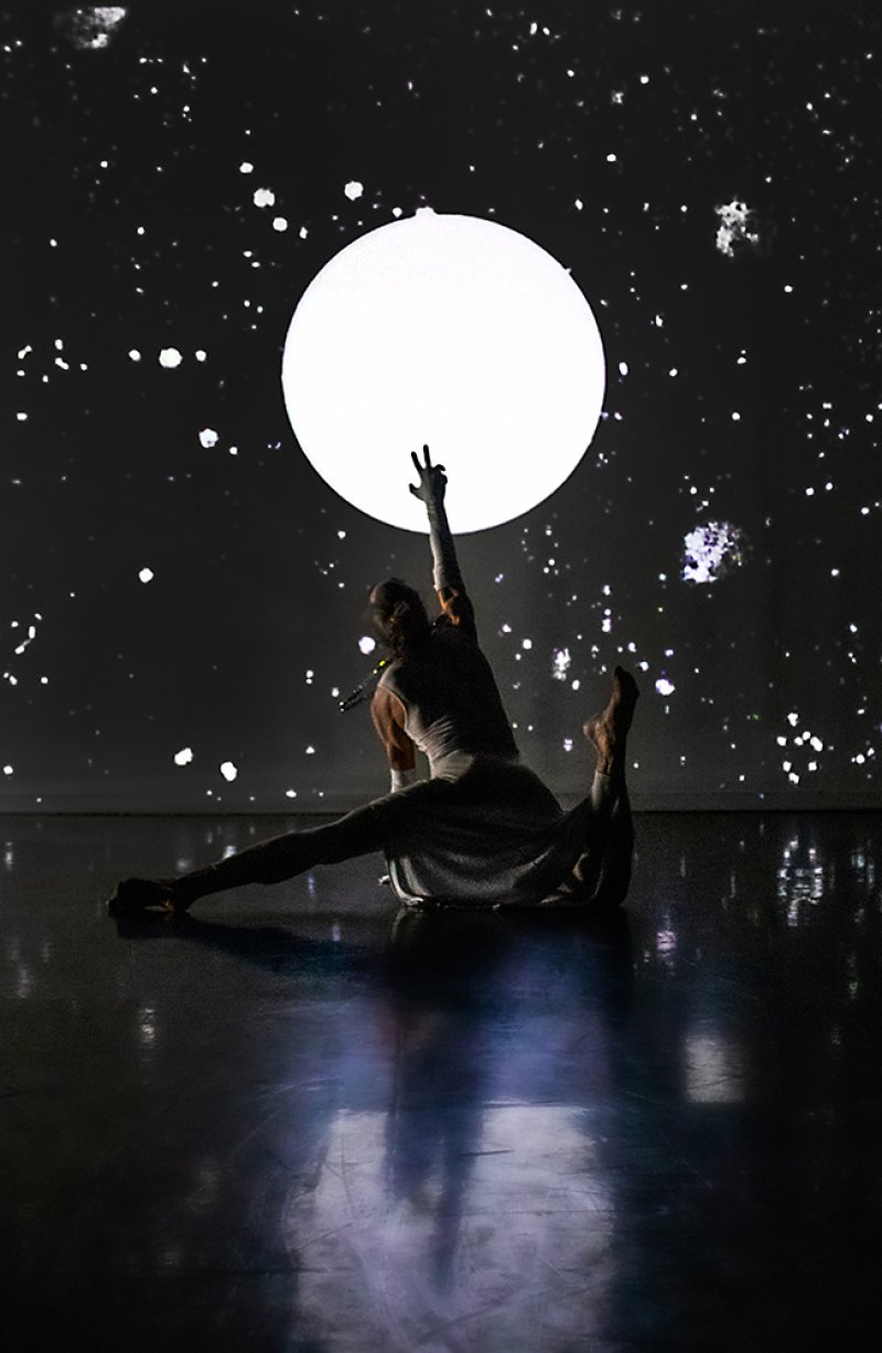 Dancer in white, silhouetted against a full moon projection and starry night sky, reaching up with one hand.