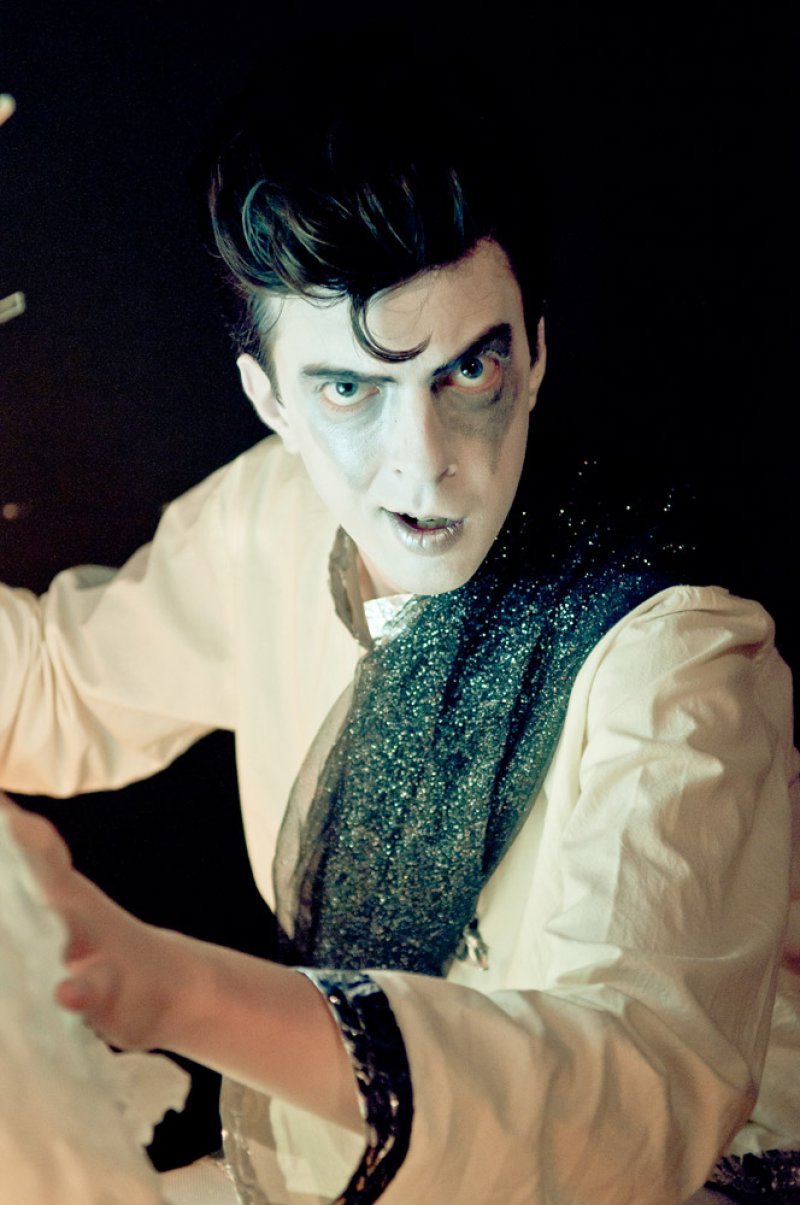 A performer in white face paint and dark eye makeup, wearing a cream shirt and a sparkly black sash, looks forward.