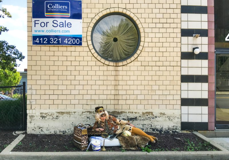 A photo shows a person with blonde hair reclining on a leopard print blanket in front of a brick building with a round window and a "For Sale" sign. The person wears patterned clothing and yellow tights.