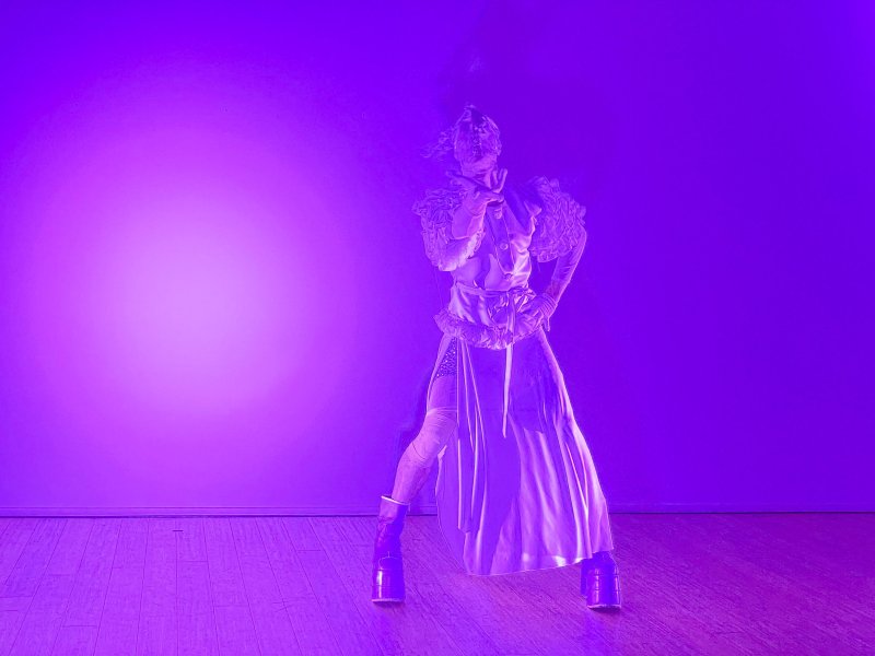 Performer in elaborate white costume with ruffled shoulders and platform boots, bathed in intense purple light, striking a pose.