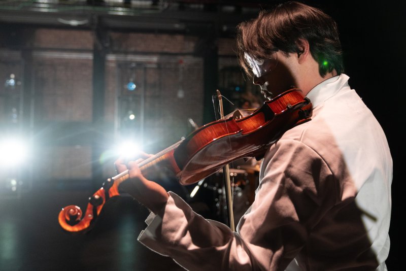 Man in white playing a violin, illuminated by bright stage lights from the left, with a dark, blurred background of stage equipment.