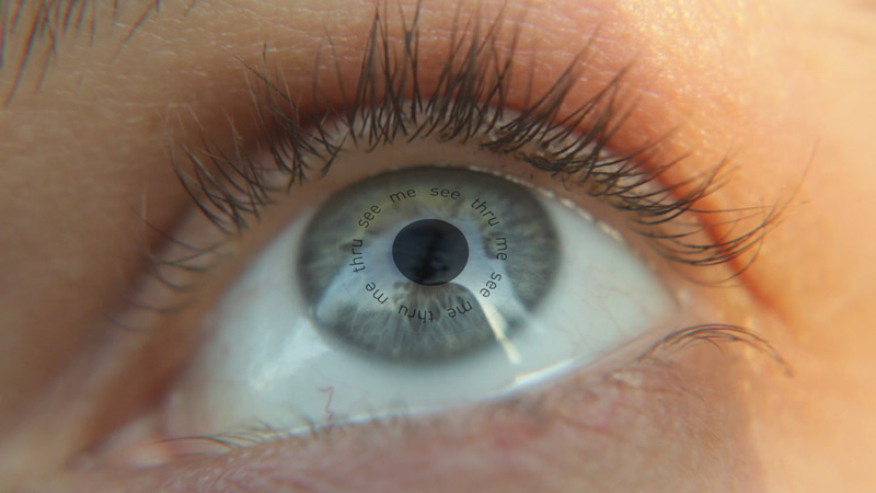 Close-up of a blue eye with "see me thru me see thru me" in a circle around the pupil, which is a dark sphere.